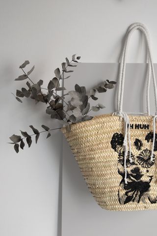 View of a raffia basket bag by Carlos Valencia featuring a black animal with flowers for eyes, the words 'UH HUH' above and white rope handles. The bag has grey foliage inside and it is pictured against a light grey background