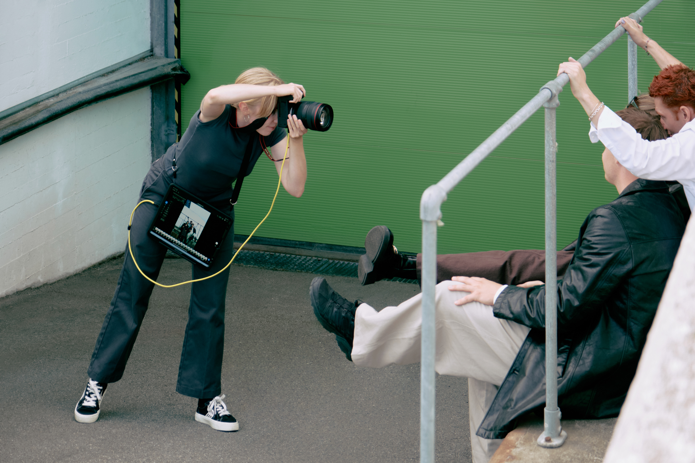 Photographer takes pictures of two people sitting on stairs with green wall background during outdoor photo shoot