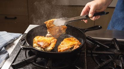 Someone turning skin-on chicken thighs in a cast iron skillet with metal tonges.
