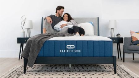 A couple laying on Bear Elite Hybrid mattress with Celliant in bedroom with black pattern rug, grey headboard and black bedside tables with lamps