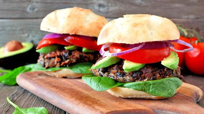 Two sweet potato and black bean burgers in buns with trimmings on wooden chopping board, a tomato and half an avocado are seen in the background