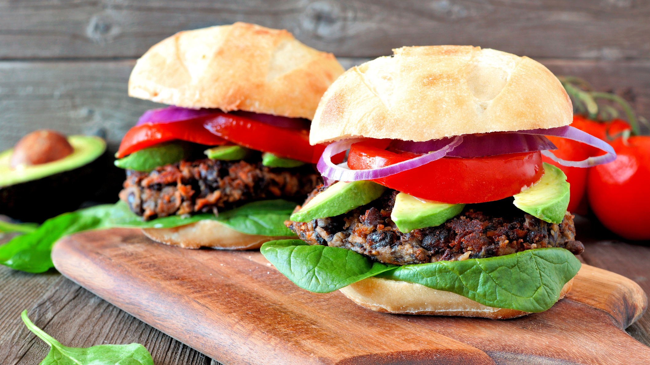 Two sweet potato and black bean burgers in buns with trimmings on wooden chopping board, a tomato and half an avocado are seen in the background