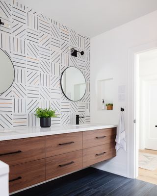 bathroom with patterned tiled walls, wall mounted wooden vanity, black floor, round mirror, wall lights, small alcove