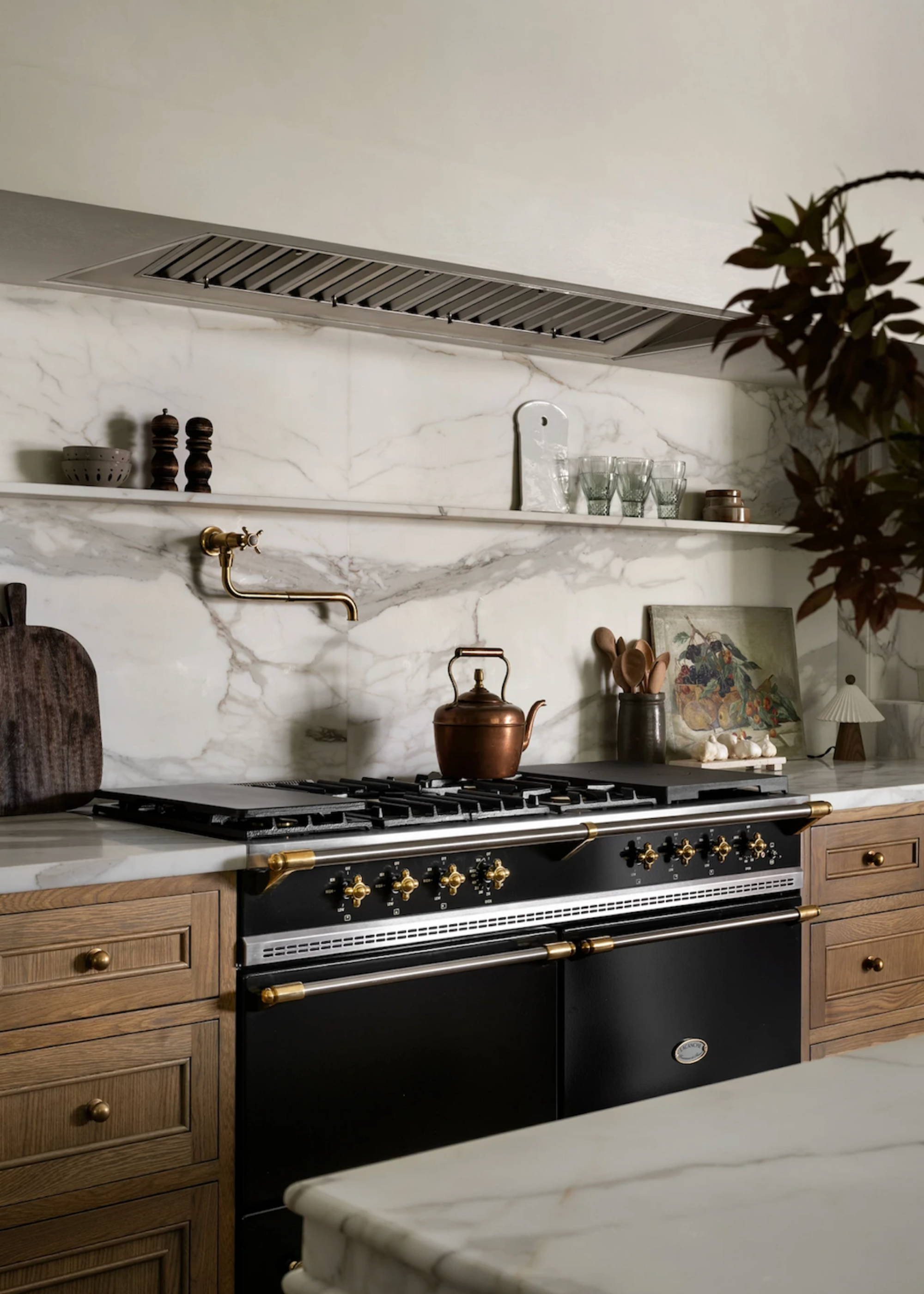 A kitchen with a slatted exhaust, a marble backsplash, a floating shelf, wood drawers, a stove with gold hardware, a brass kettle, a filter tap, a chopping board, a glass of wood utensils, and some glassware