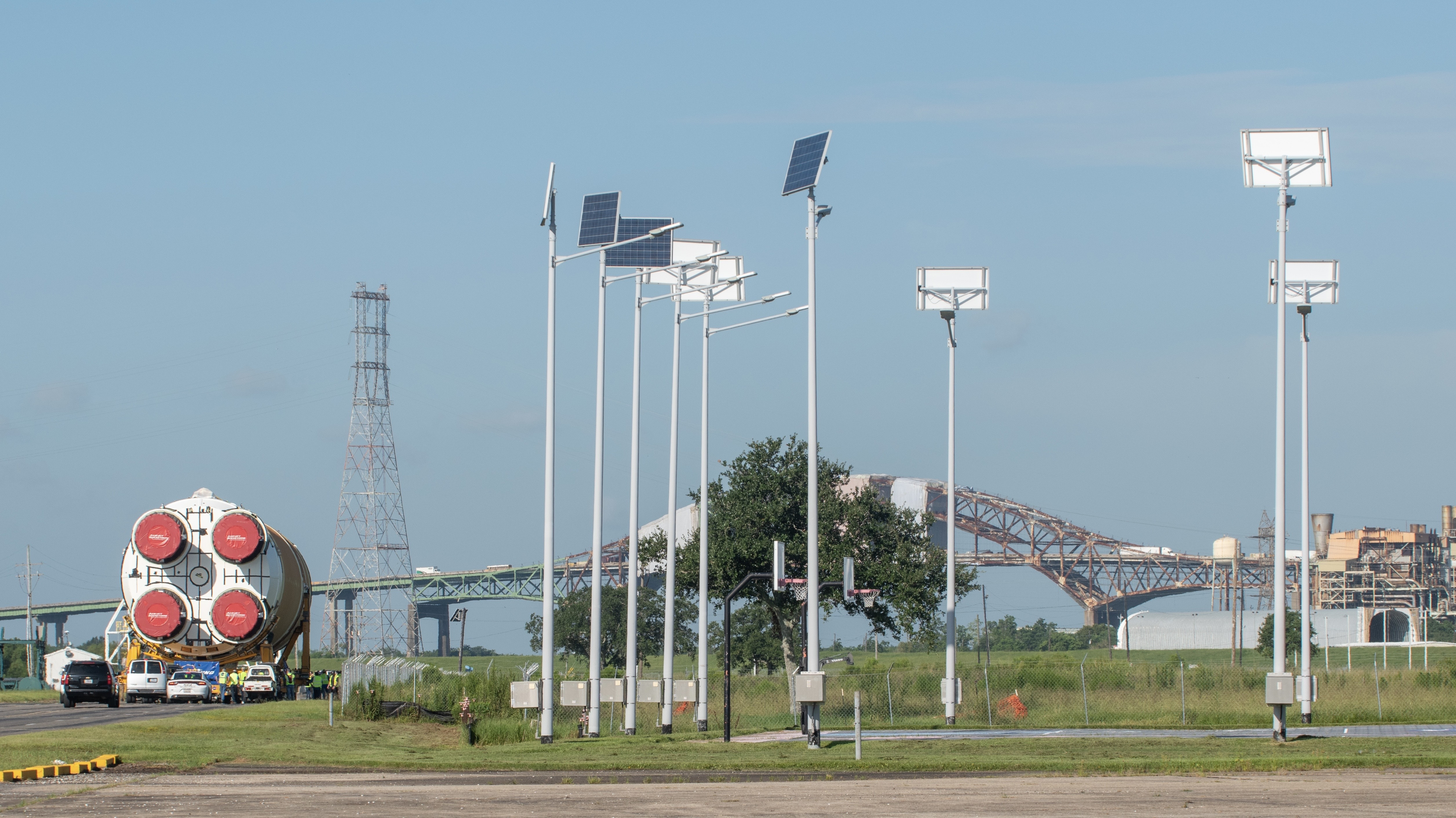 The tail of a rocket booster with its four engine caps covered in red tarpaulin can be seen moving slowly towards a tubular barge in the distance to the right.