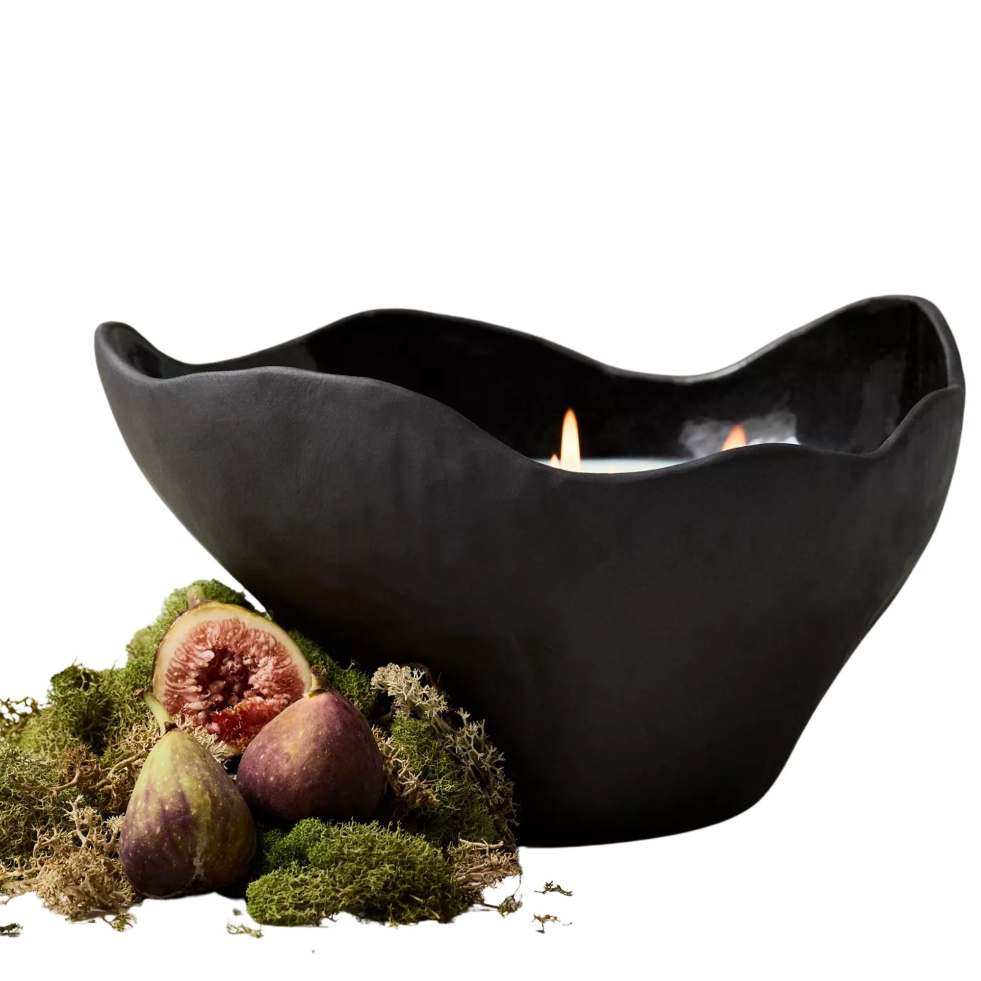 A lit fig candle in a waved black ceramic vessel with fresh figs and moss in the foreground.