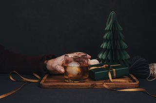 A person holding a glass of brandy with ice, next to small, green gift box and a paper Christmas tree decoration, on top of a wooden board.