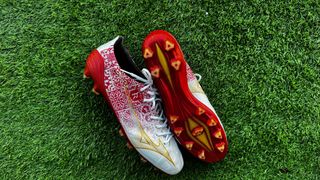 Red and white football boots with a flash of gold on the side are together on green artificial grass. Mizuno Alpha Made in Japan football boots