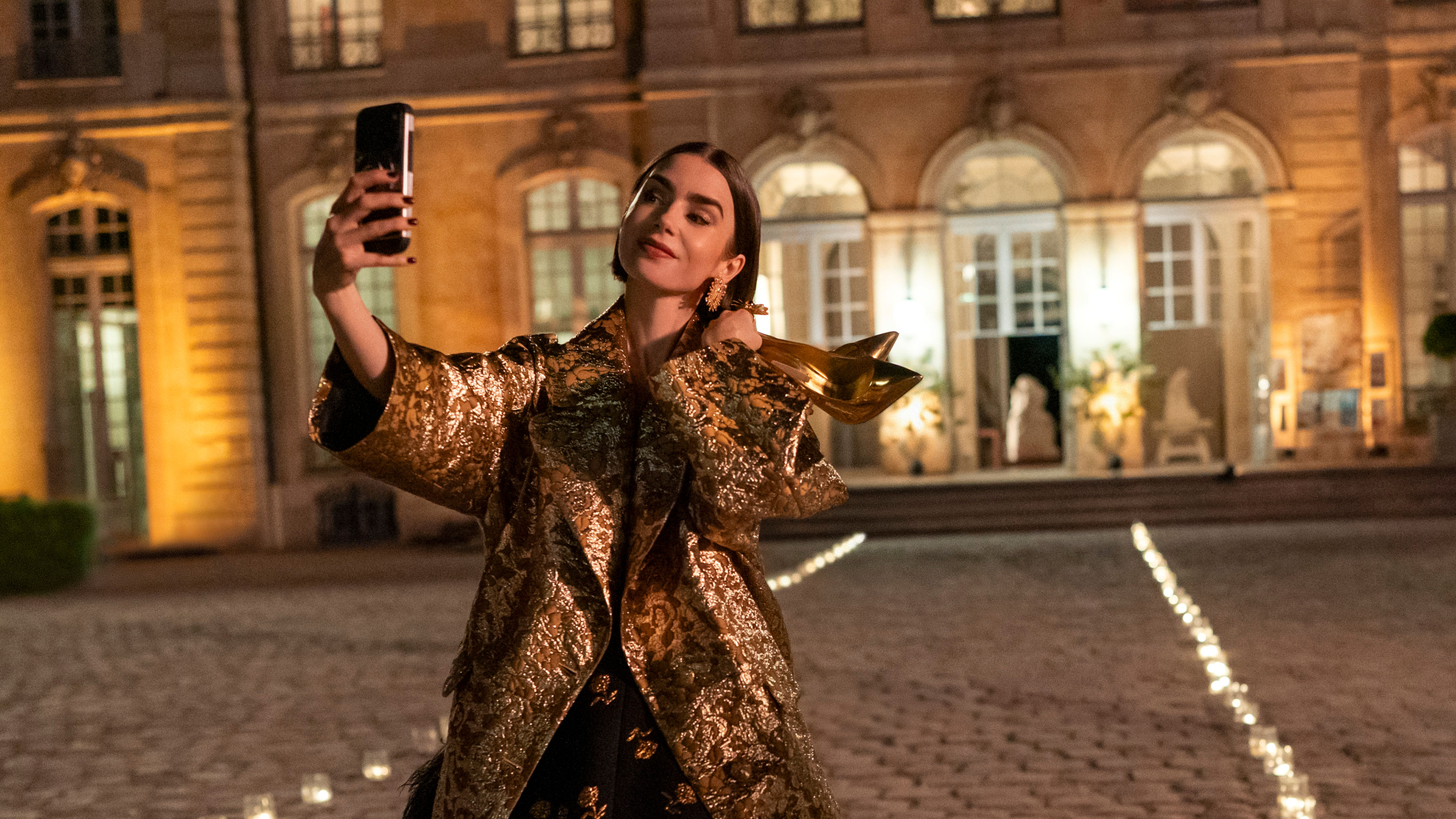 lily collins as emily taking a selfie outside of a european building at night while wearing a gold coat and holding a pair of gold heels in emily in paris season 6