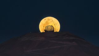 an orange moon rises from behind the top of a dark hill, shining through the scaffolding frame of a domed observatory.