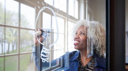 A smiling woman draws a lightbulb on a window.