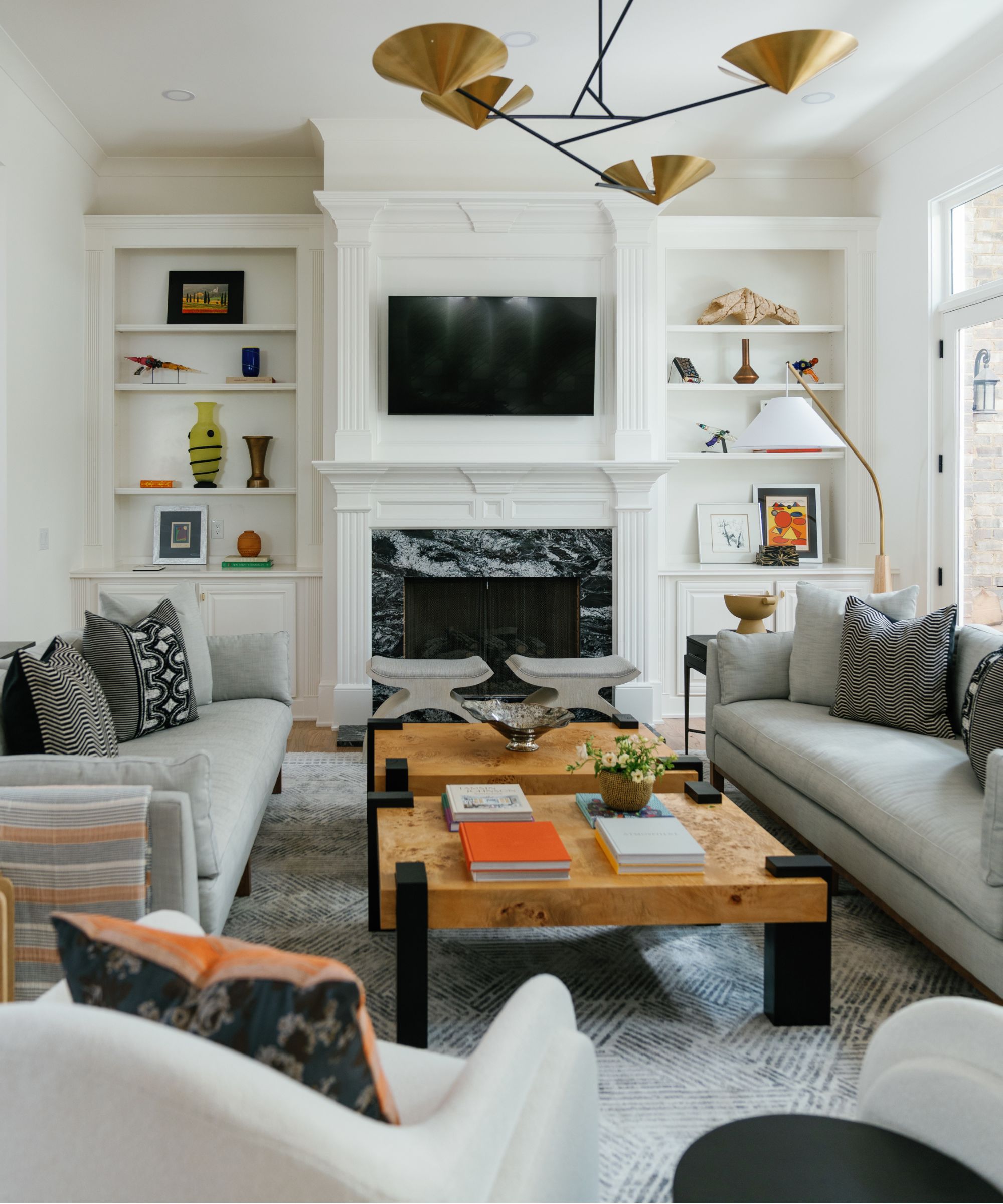 A transitional living room with white built-in shelves holding vases, trinkets, and pictures, a marble fireplace with a TV above, a burl wood low coffee table, two pale grey sofas, and an armchair.