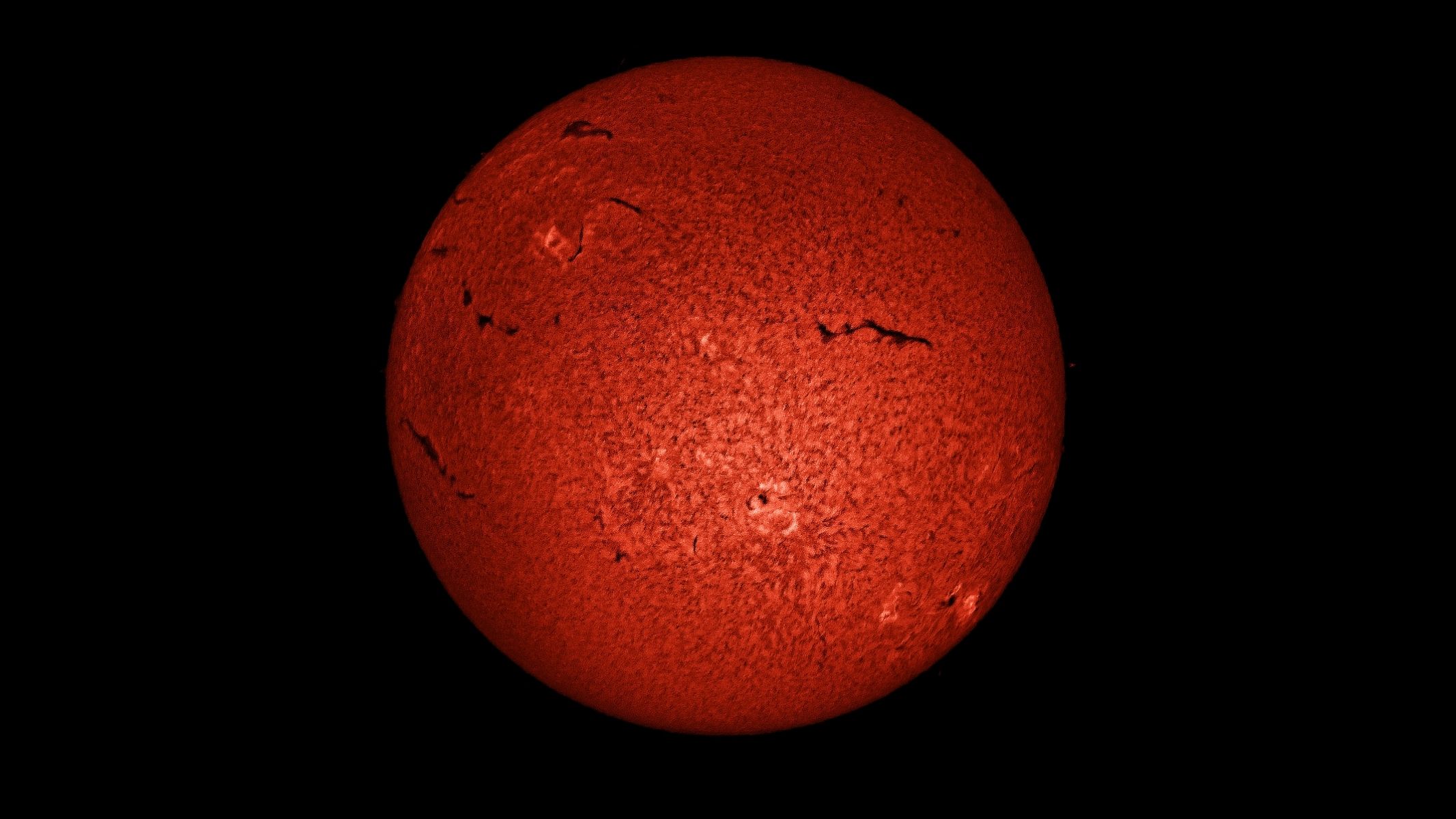 The solar disk in deep orange, showing a textured surface and dark spots, as imaged using the Sky-Watcher Heliostar 76mm H-alpha solar telescope.