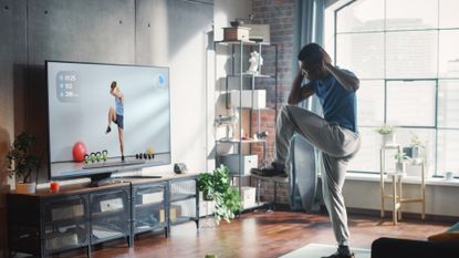 man wearing joggers and tshirt performing a standing elbow to knee move in front of a large tv screen in a home setting. 