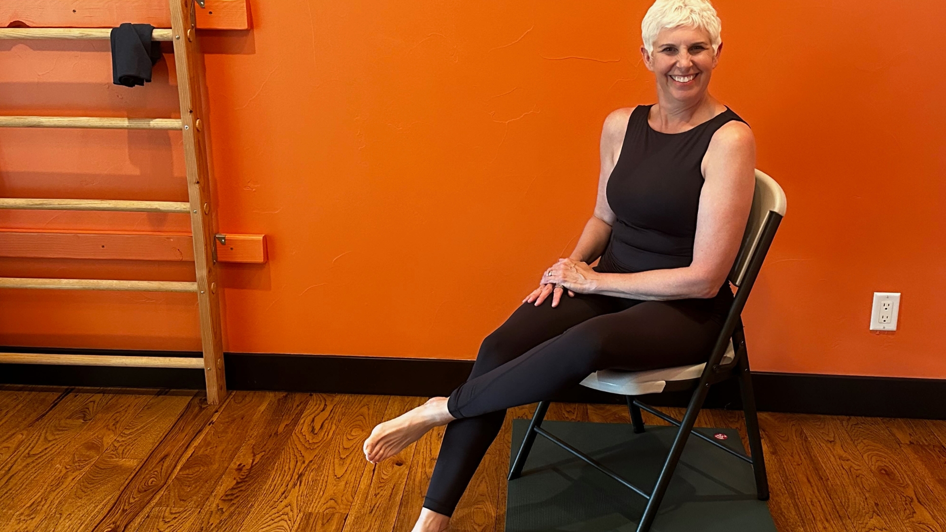 woman on a chair with one ankle over her other thigh. there's an orange wall behind her.