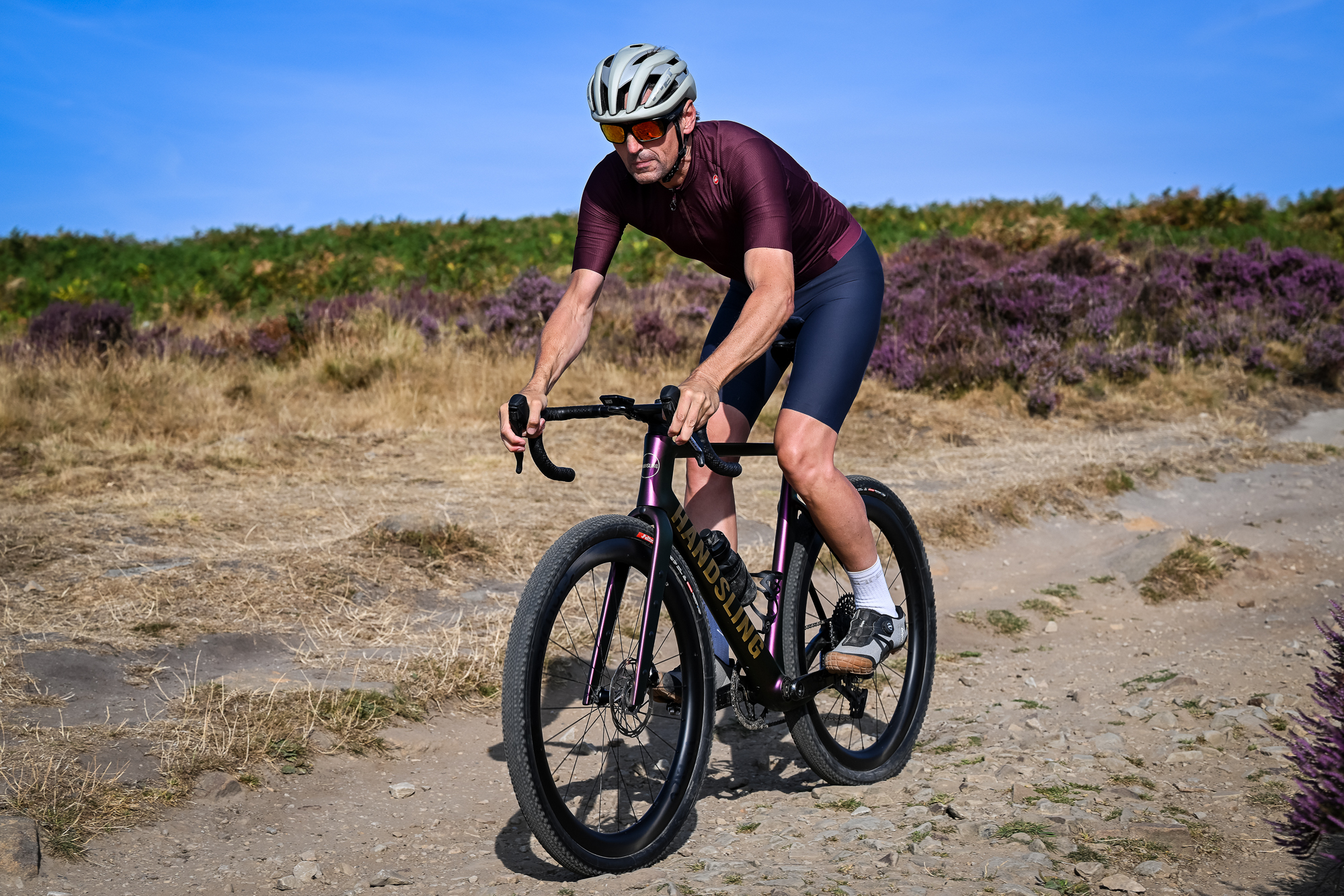 Man riding a purple gravel bike down a sandy trail wearing grey shoes, blue shorts, a purple top and a beige helmet.
