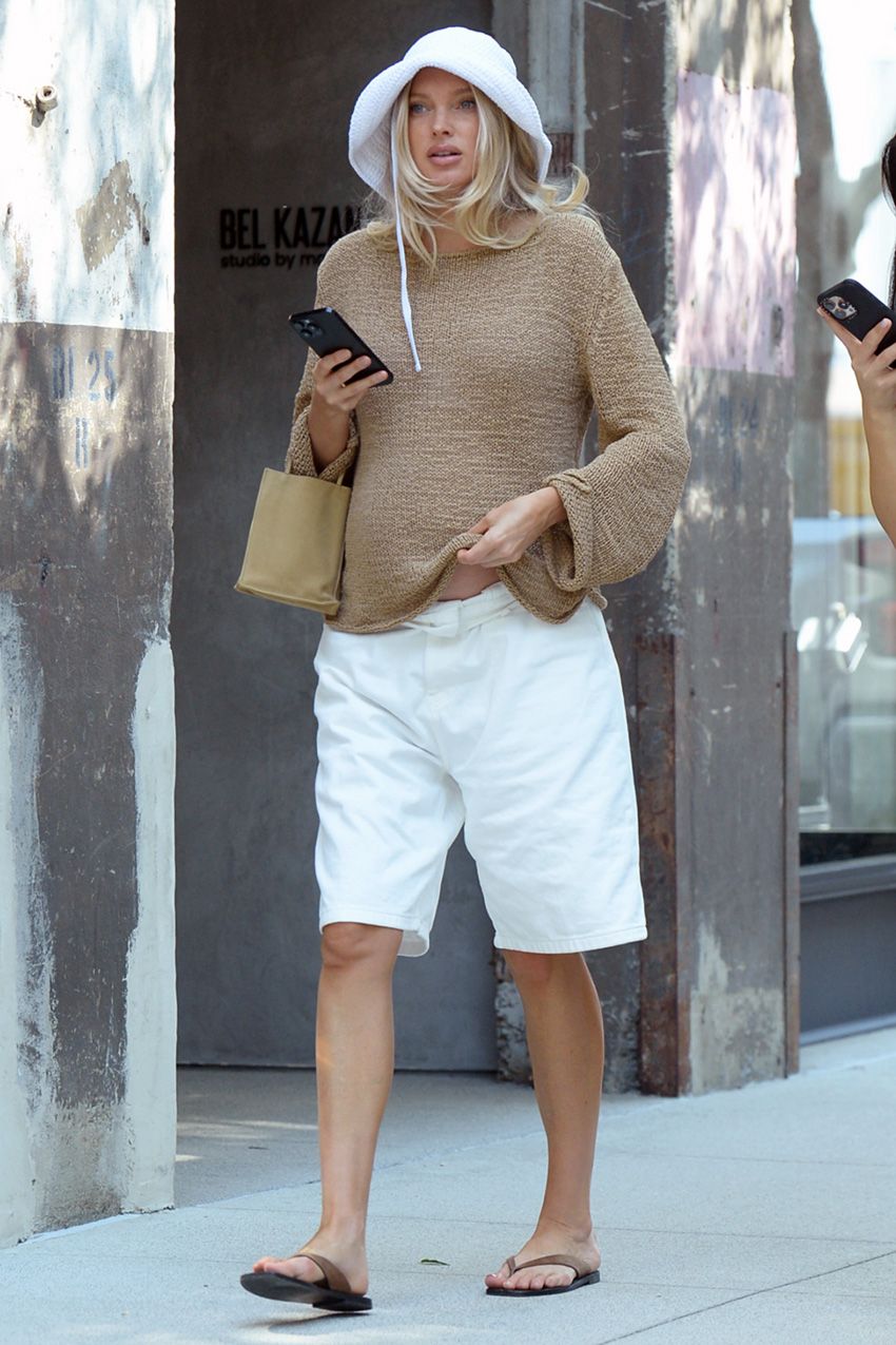 Elsa Hosk wearing a white bucket hat, tan summer sweater, white bermuda shorts, flip-flops, and a The Row Cecily Mini Tote Bag