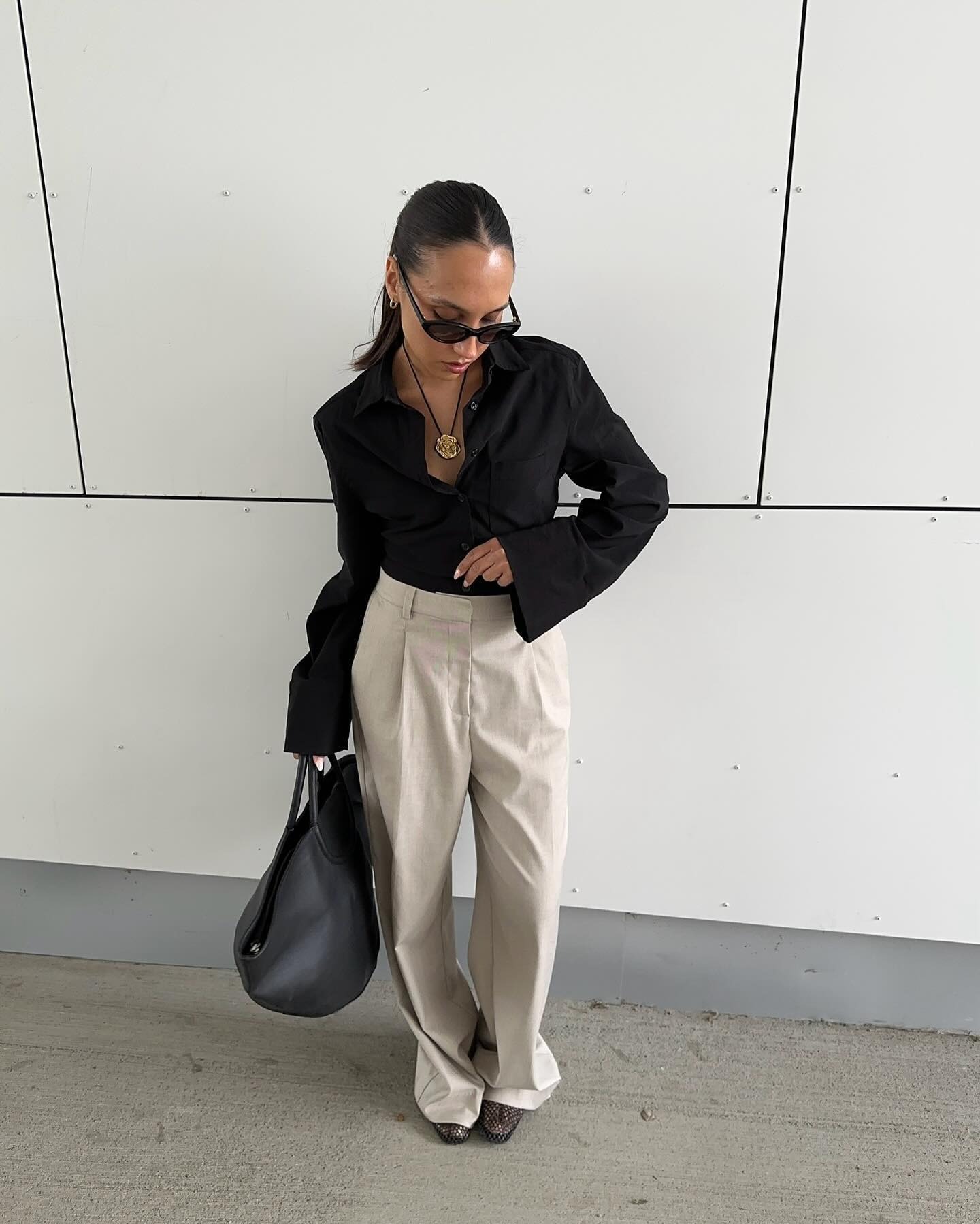 Influencer wears beige trousers tucked into a black blouse. She stand outside and holds a large black handbag and styles it with brown shoes and a gold cord necklace.