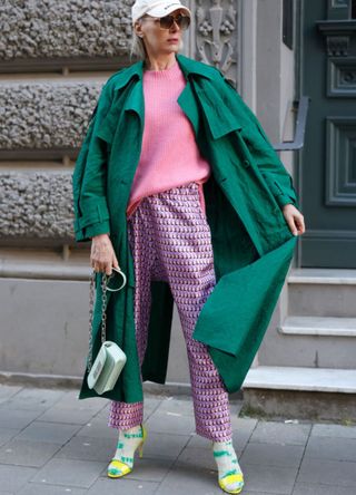 Model and Influencer Petra van Bremen wearing a pink knitted pullover by Van Kukil, a dark green coat by Christian Wijnants, pink pants with abstract pattern by Odeh, green and beige tie-dye cashmere socks by mell-o, sunglasses by Spektre Eyewear, a cream colored cap by Balenciaga, yellow high heel sandals by H&M and a pastel mint green bag by Balenciaga