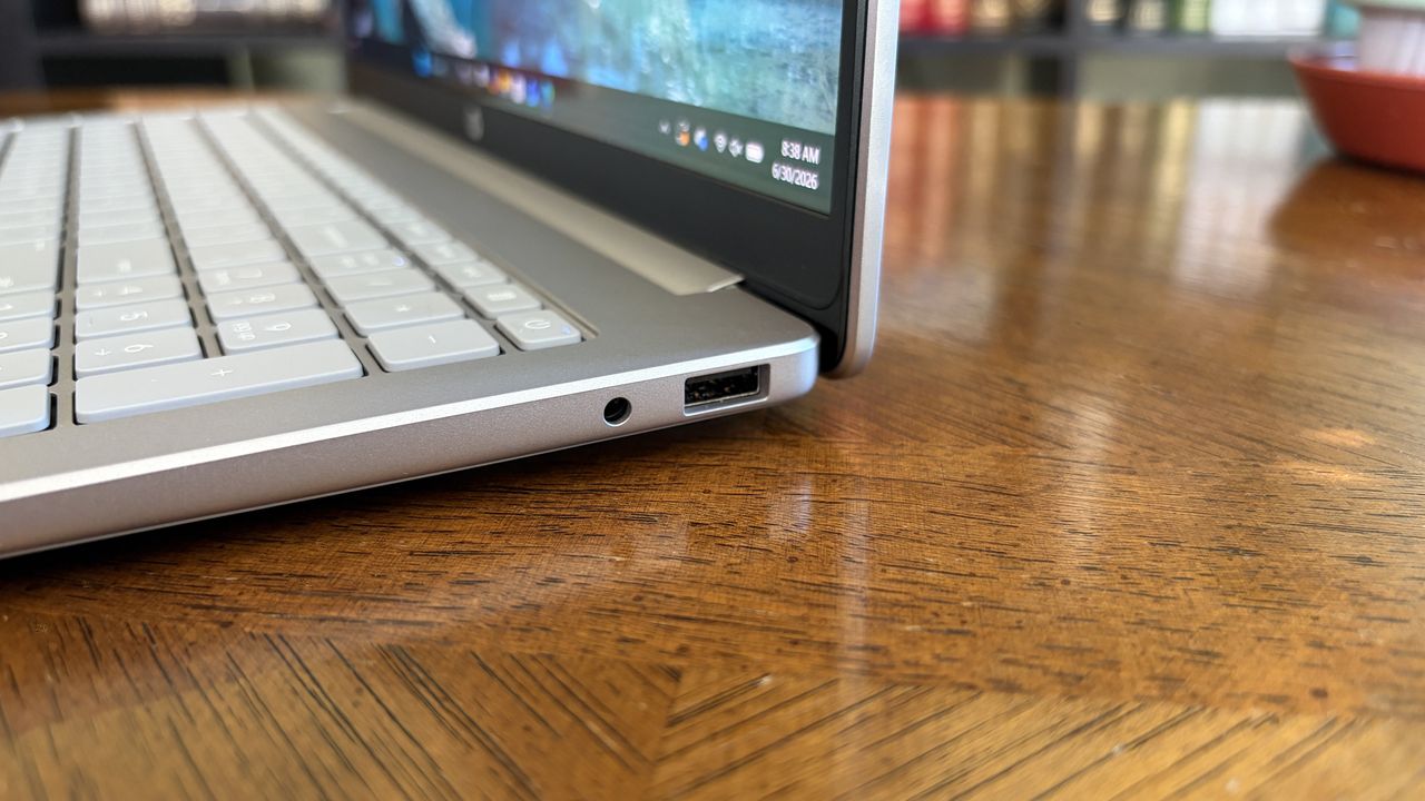 A silver OmniBook 3 laptop viewed from the side, displaying several ports, sits on a wooden table with a book-filled background.