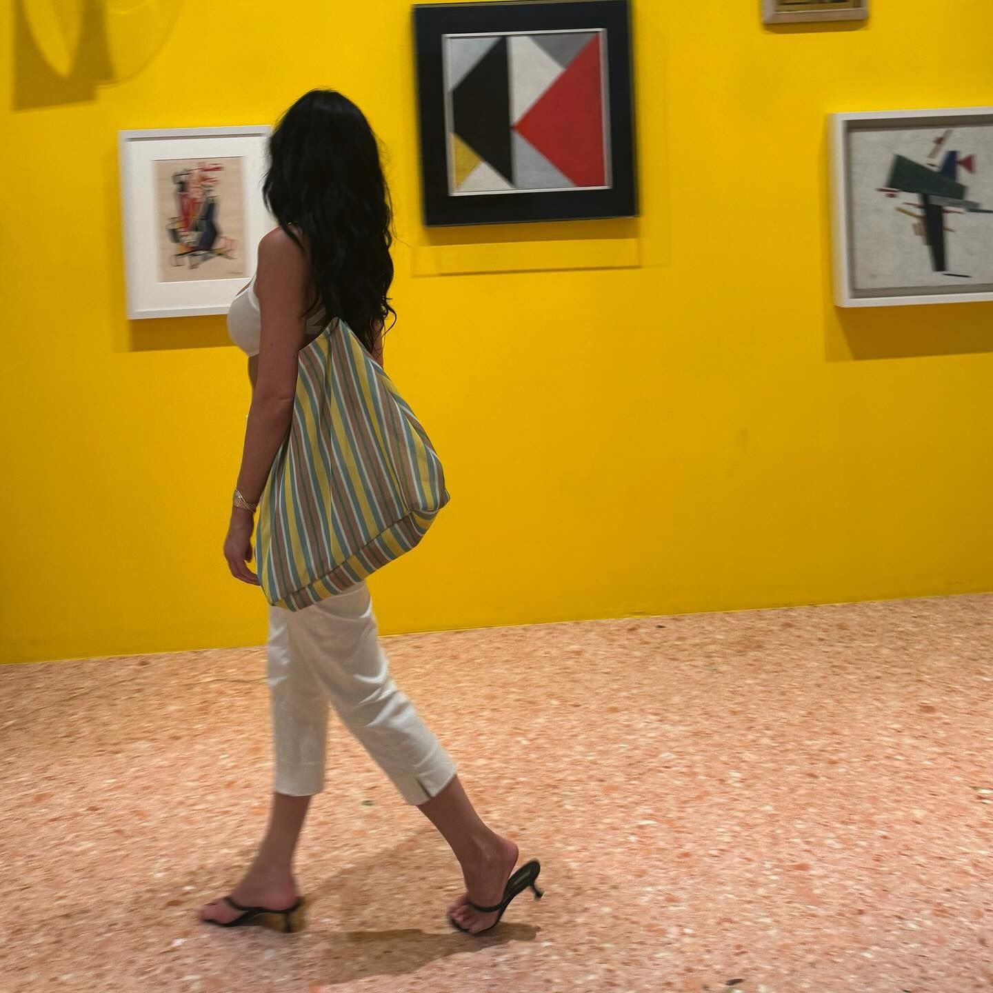 Kylie Jenner wearing white capris, white tank top, black sandals, and The Row mesh market tote in art gallery.