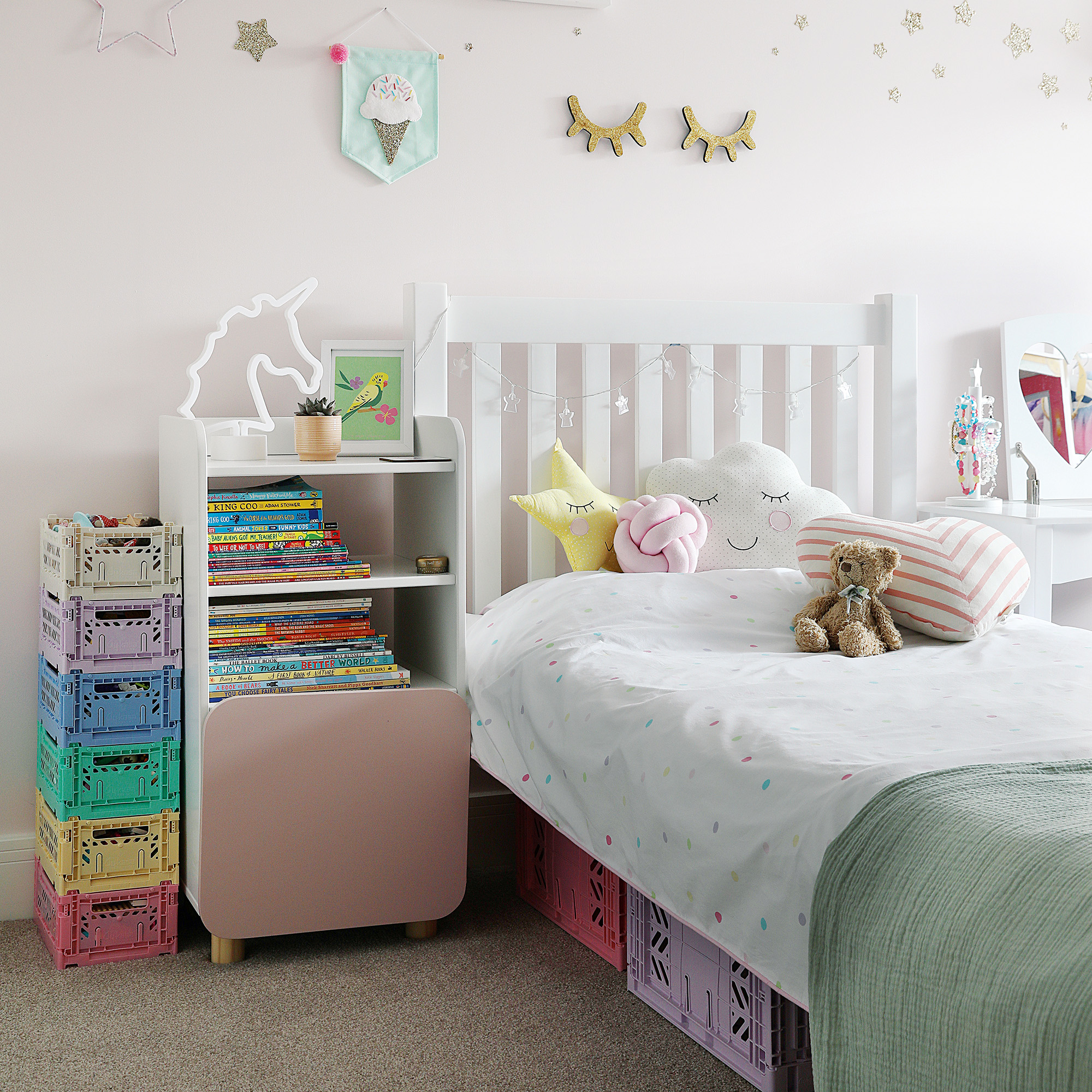 girls bedroom with pale pink walls white wooden bed frame with character cushions, white and pink bedside table filled with books and a stack of multicoloured mini plastic crates