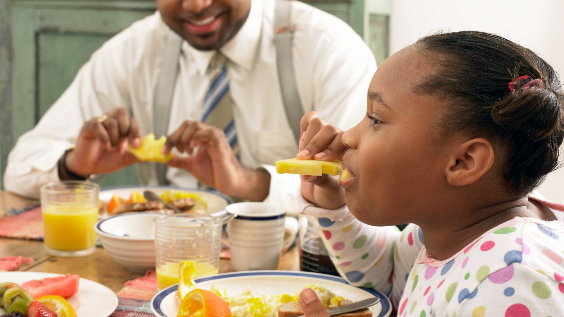 A young girl and her father eating pineapple at the breakfast table.