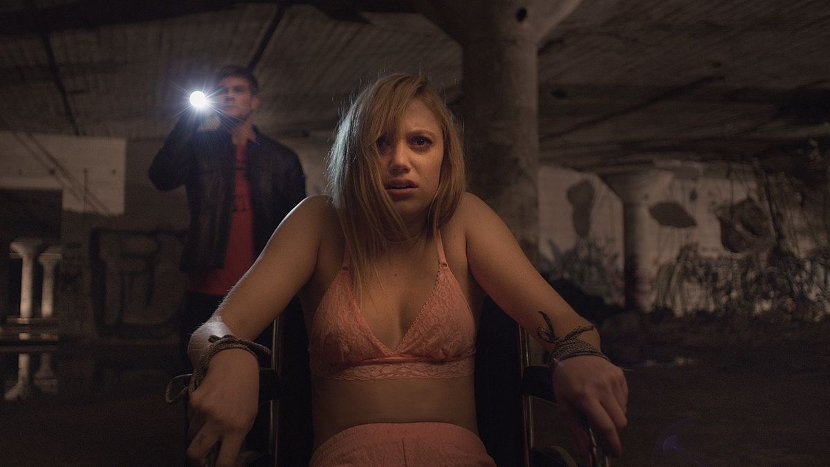 Jay sits in a chair looking scared as Hugh watches on in It Follows