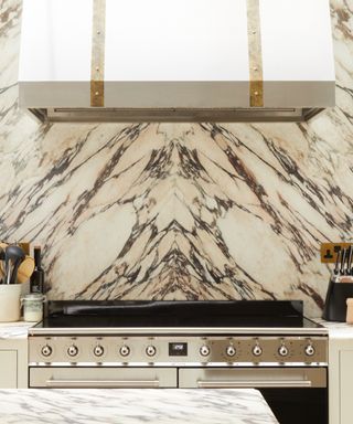 Marble and white kitchen with white cooker hood with brass detailing, chrome and black oven, marble backsplash, marble countertops, utensils,