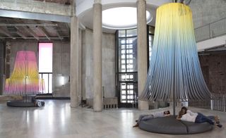 Lamp-like art installations made of ribbons in pink, yellow, and blues shades, are set throughout the room with high ceilings in gray tones. There is a seating below the art installations, with people on it.