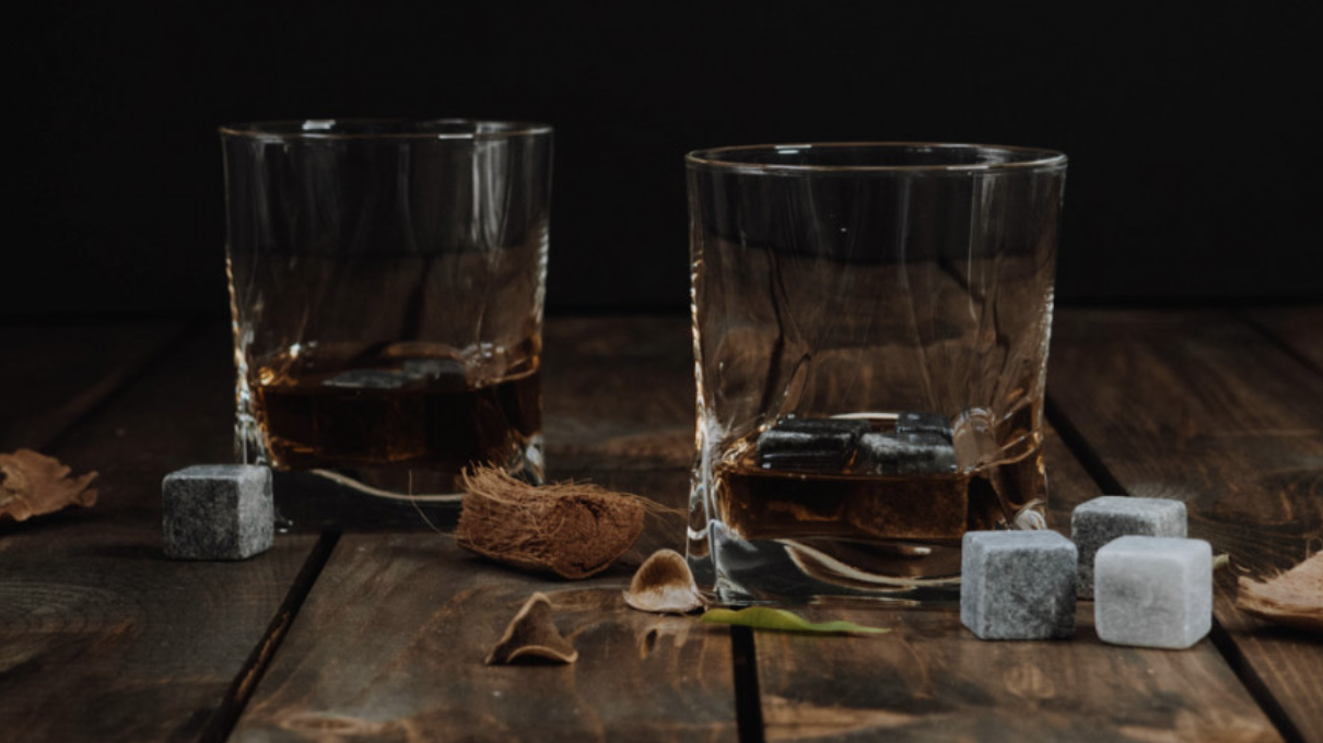 the image depicts two whisky tumblers on dark wooden table. Each glass has a small measure of brown whisky in it with a couple of ice cubes