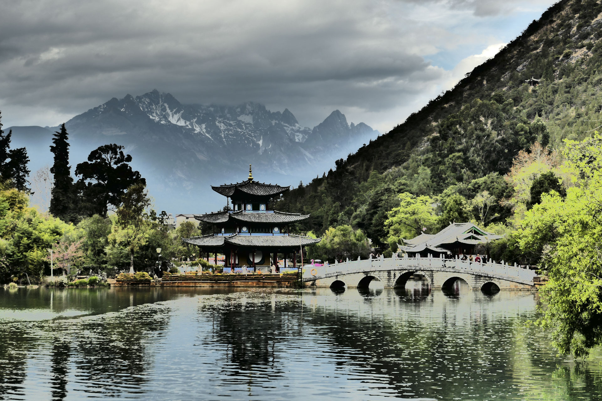 A Chinese temple by the side of a lake with an arched bridge leading up to it and both are reflected in the still water, in front of a snow covered mountain. A creative filter is applied to the image