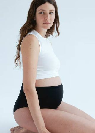 A pregnant model wearing a white tank top and black maternity bikini bottoms from H&M. 