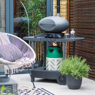 Portable gas bbq on a patio with light grey outdoor tiles and a wicker outdoor seat
