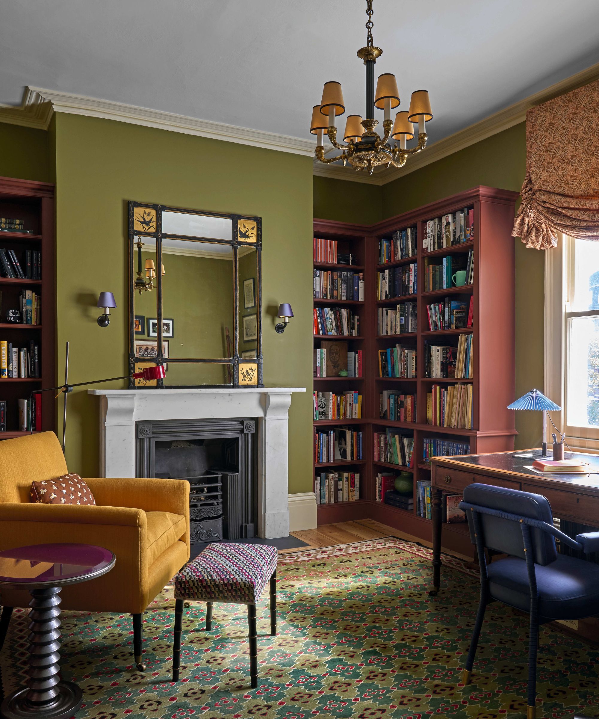 A classic study with olive green walls, built-in reddish-brown bookshelves, and a white marble fireplace. A mustard yellow armchair and a patterned rug add warmth to the room.