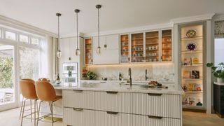 pale coloured kitchen with large island, tan leather bar stools, three glass and brass pendant lights above island and back wall of cabinets, some glass fronted, others open and some with full doors, plus worktop with kitchen sink