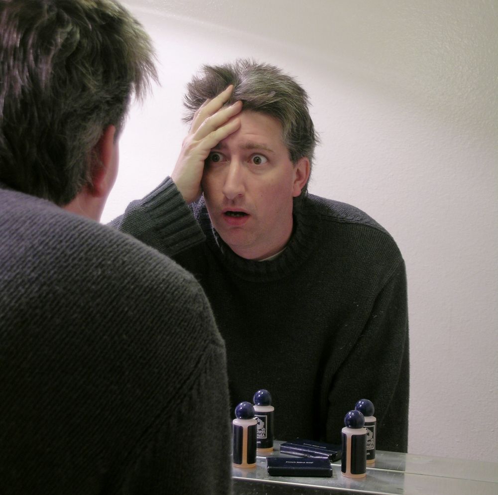 Man in a dark sweater is looking in the mirror of a hotel room with a shocked look on his face. 