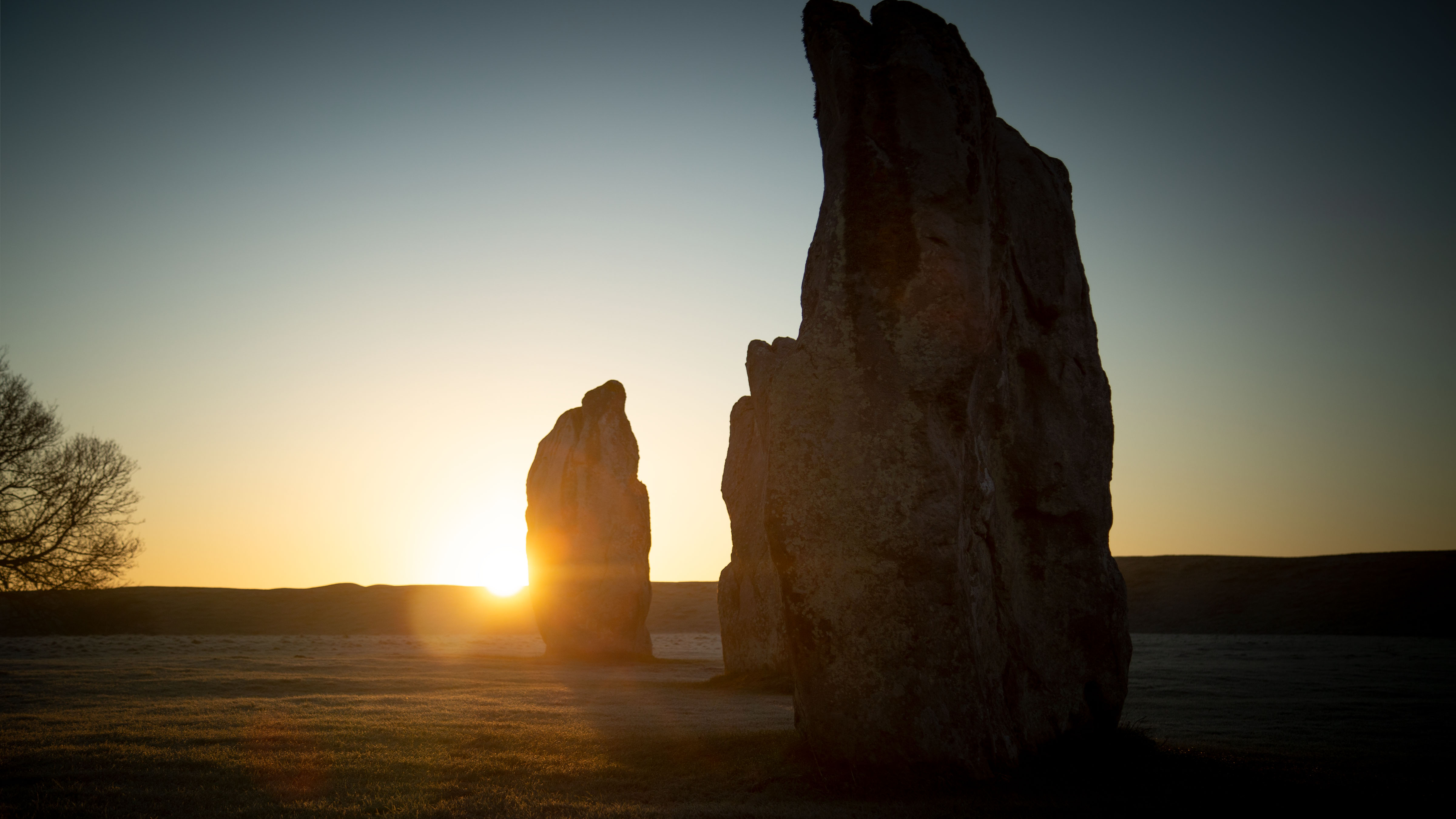 the sun rises behind a stone towards the left of the picture. taller stones stand in front in shadow