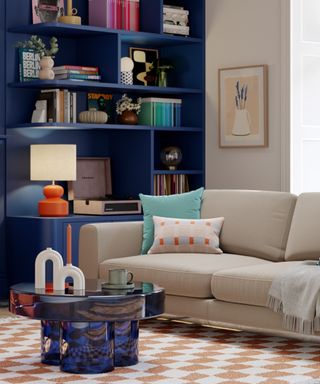 A living room with a dark navy blue bookcase with books and decor on it, a cream wall and a cream couch to the right with a dark blue table and white and brown checkered rug underneath it
