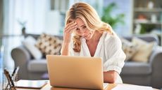 A woman stares at her laptop at her table at home, looking like she has a headache.