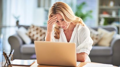 A woman stares at her laptop at her table at home, looking like she has a headache.