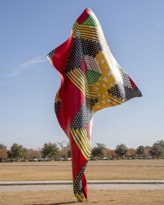 A wind-swept sculpture made of a textile-like material in red, yellow, blue, pink, green, and white tones features traditional African patterns while standing tall in a sunny garden.