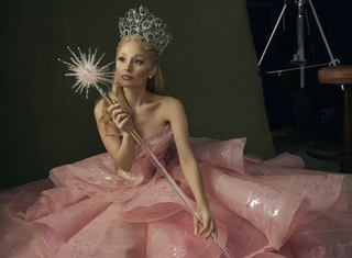 Ariana Grande sits on a set for a promotional Wicked photoshoot wearing a pink strapless dress and tiara for her character Glinda