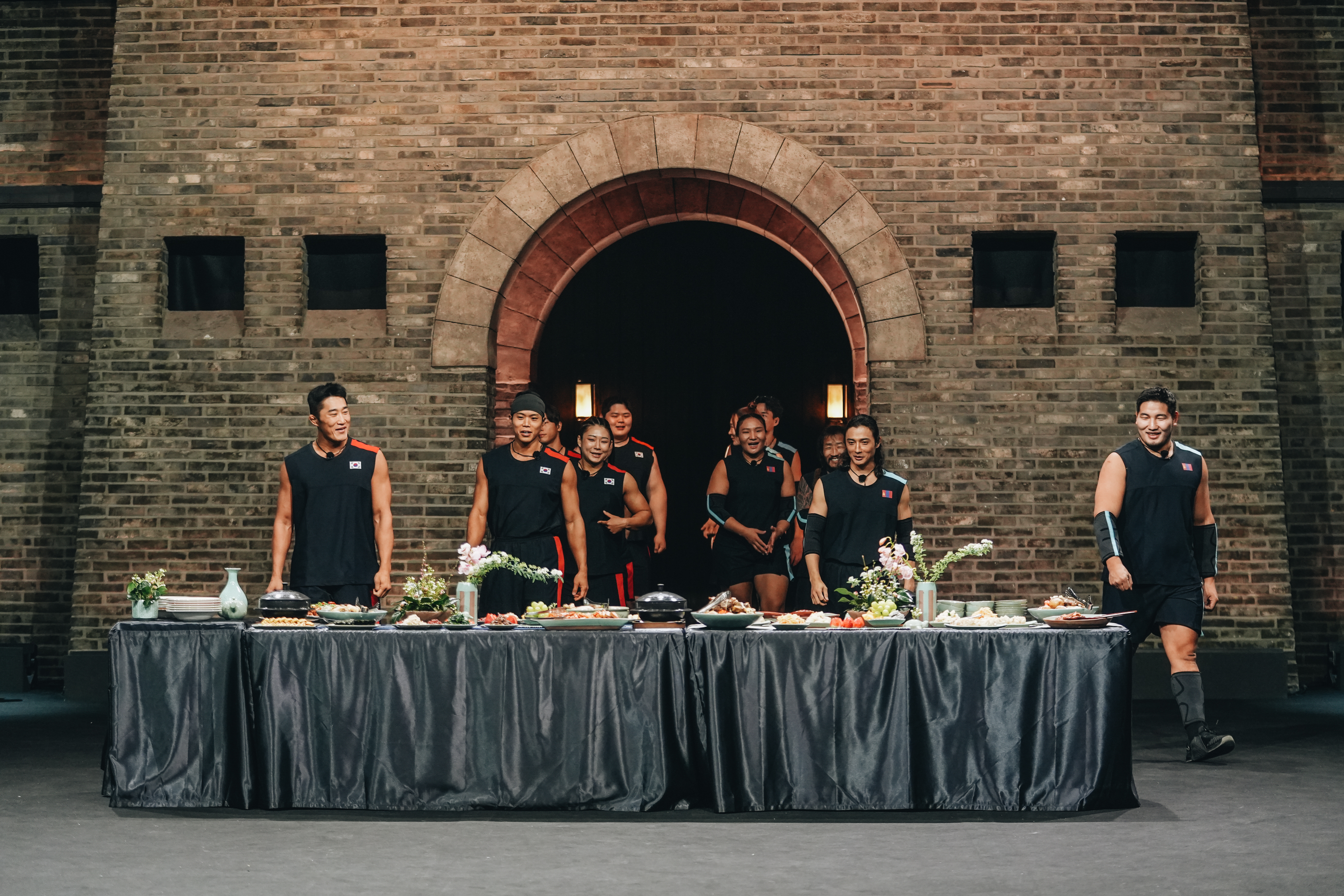 A group of people (l-r Kim Dong-hyun, Amotti, Choi Seung-yeon, Kim Min-jae, Adiyasuren Amarsaikhan, Lkhagva-Ochir Erdene-Ochir, and Orkhonbayar Bayarsaikhan) enter a chamber with a table of food, in Netflix&#039;s &#039;Physical: Asia.&#039;