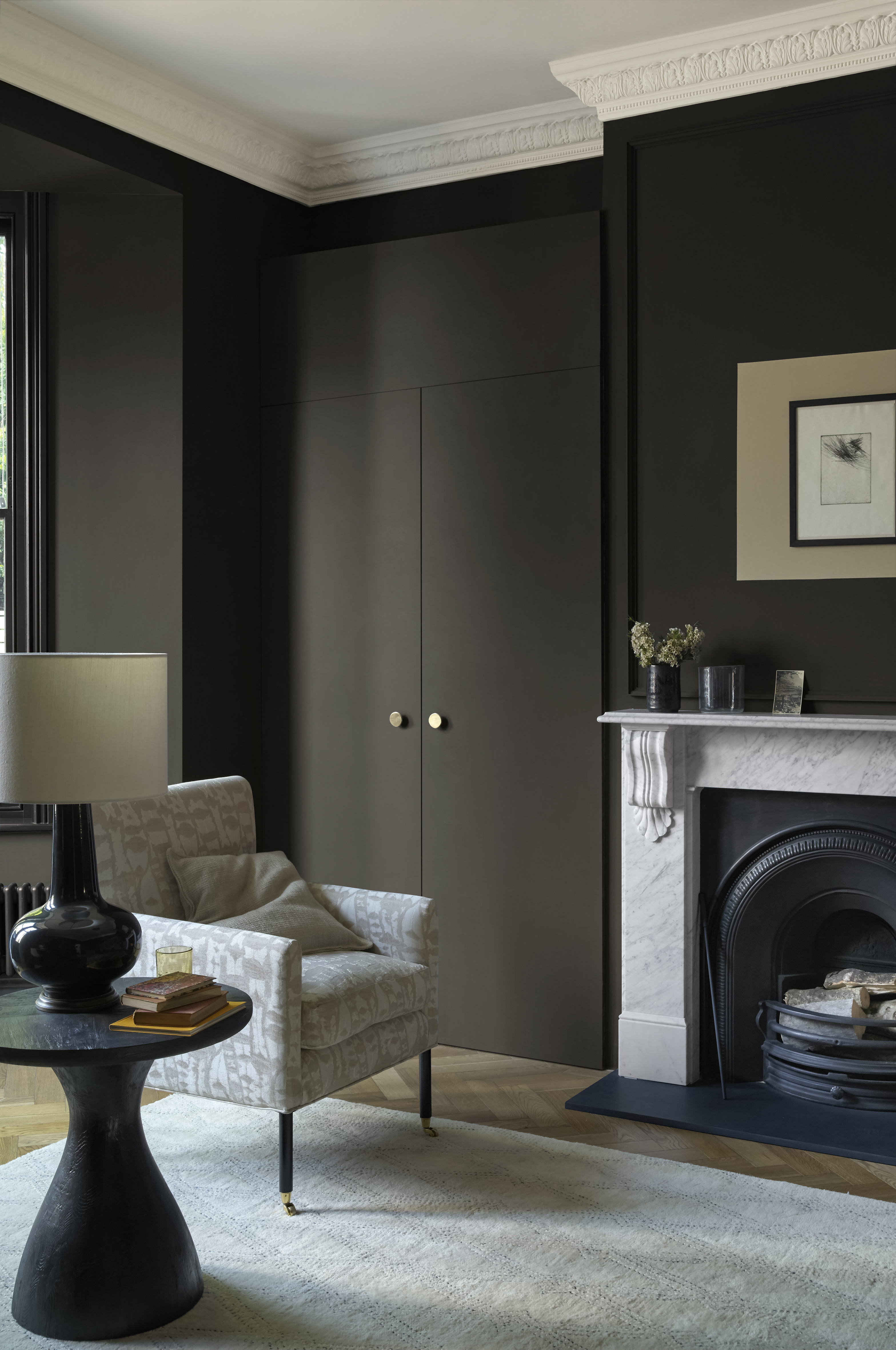 Living room painted in dark brown with fireplace and armchair in pale stone, a cream rug and black accessories.