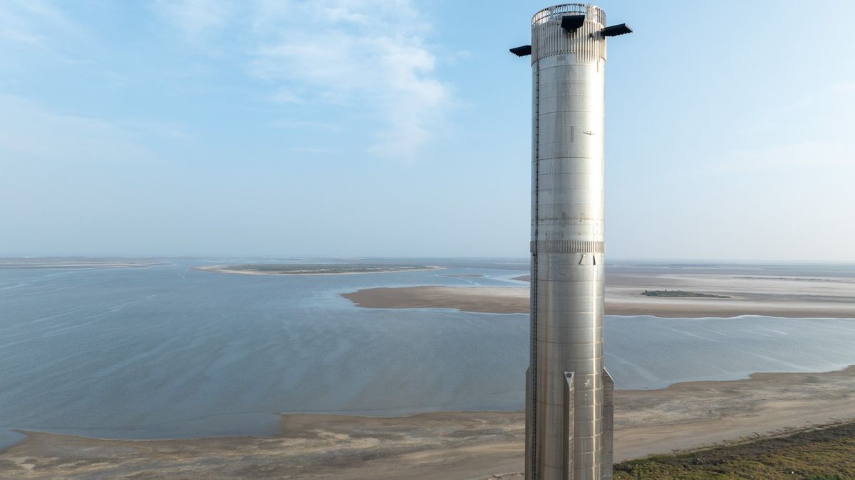 a very tall, silver rocket rolls along on a truck on a road next to the ocean