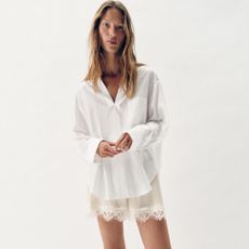 Zara model with wavy hair posing in a a chic summer 2026 outfit wearing an oversize white button-down shirt and beige lace-trim shorts