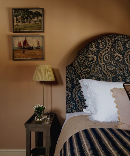 Bedroom with earthy pink walls, a blue floral headboard, striped wool blanket, framed art and a pleated bedside lamp.
