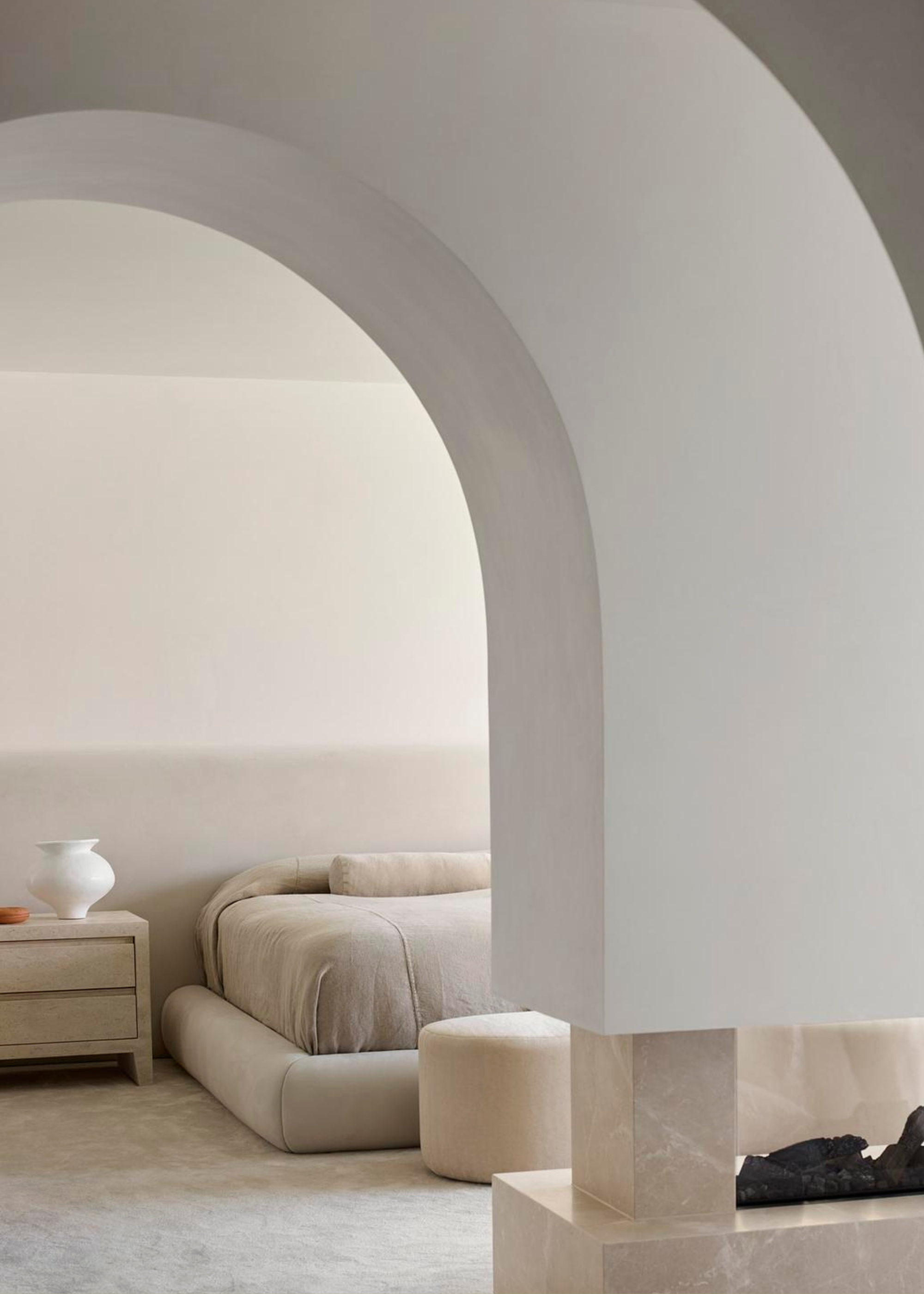 A white bedroom with arched doorways, a low bed with a side table and a pouf