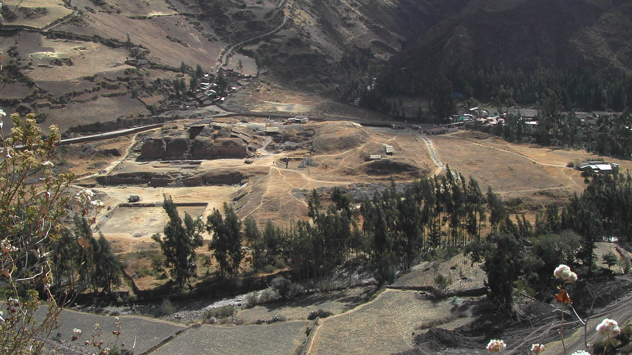 View of Chav&iacute;n archaeological site in Peru. The ground is brown with scrubby trees and mountains rising in the background.